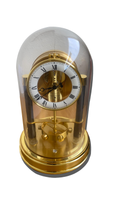 Pendule de table sous cloche en verre, boîtier doré, cadran à chiffres romains et mécanisme visible, posée sur un socle